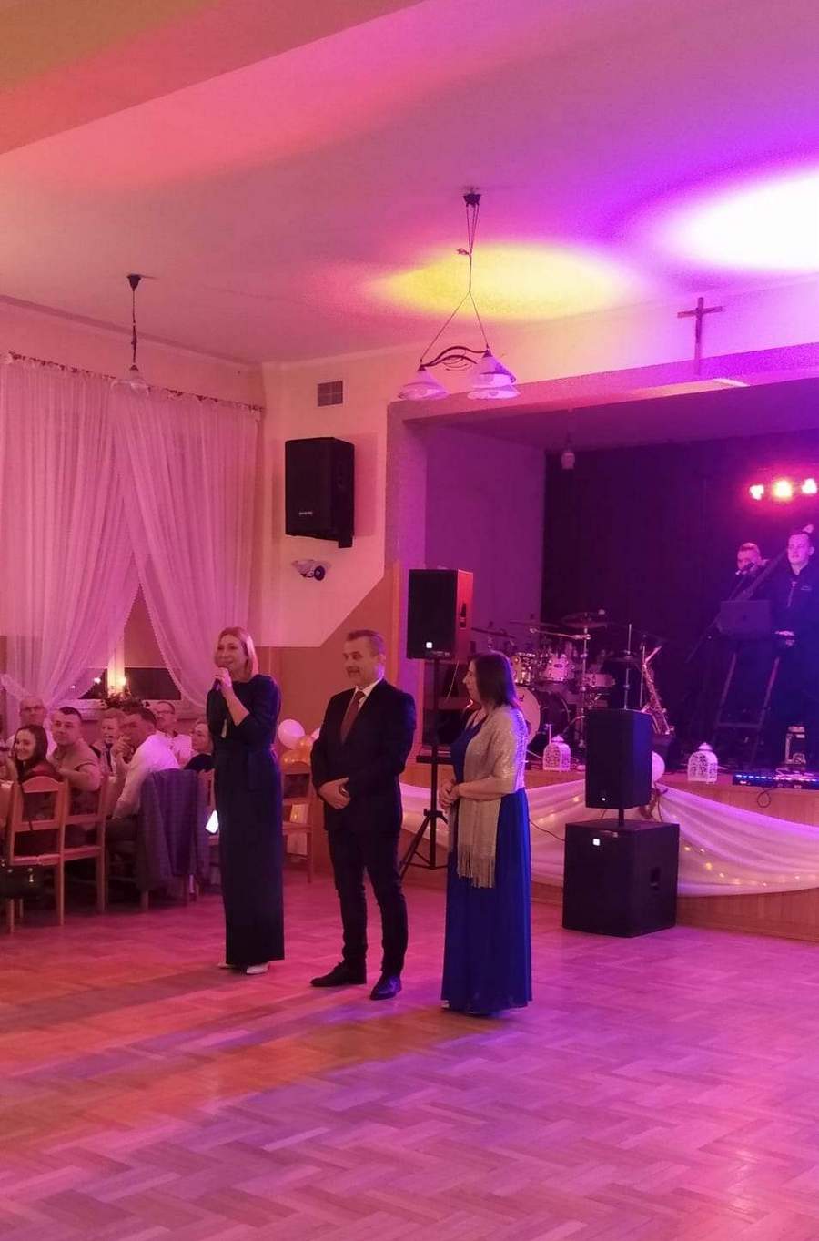Sala bankietowa oświetlona różowo-czerwonym kolorem. Na pierwszym planie trzy osoby, dwie kobity i&nbsp;mężczyzna w&nbsp;środku.