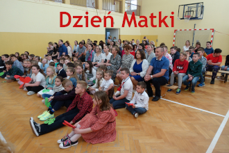 Dzieci siedzą na podłodze w&nbsp;sali gimnastycznej. Za nimi ich rodzice. Wszyscy patrzą przed siebie. Na zdjęciu napis dzień matki.