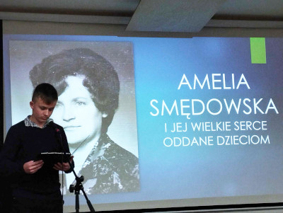 Uczeń czyta przez mikrofon. W tle wizerunek i napis Amelia Smędowska i jej wielkie serce oddane dzieciom