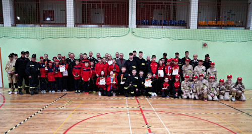 Uczestnicy Gminnych Zawodów Sportowo-Pożarniczych pozują do zdjęcia wraz z organizatorami. W ręku trzymają zdobyte puchary i dyplomy. Pozują we wnętrzu sali gimnastycznej Szkoły Podstawowej w Kwiatonowicach.