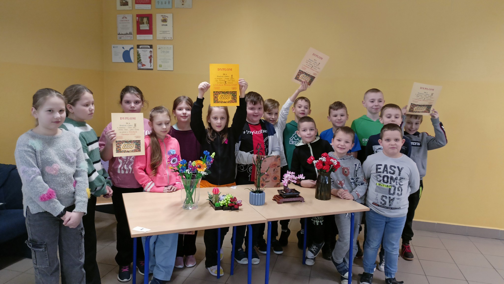 Uczniowie którzy brali udział w szkolnym konkursie LEGO trzymają zwycięskie dyplomy i stoją wokół stołu na krórtym znajdują się nagrodzone prace - kwiaty LEGO