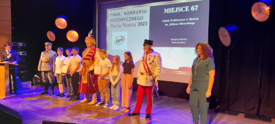 Uczniowie z dyplomami na scenie. Wśród nich troje mężczyzn przebranych w historyczne stroje. Za nimi dekoracje.