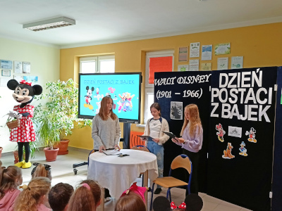 Uczniowie na dniu postaci z bajek. Trzy uczennice prezentują coś pozostałym uczniom.