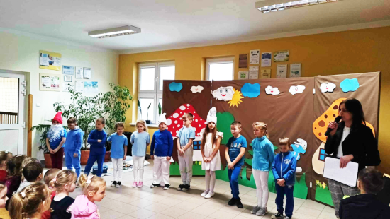 Dzieci biorące udział w przedstawieniu stoją na tle dekoracji. Obok nich z mikrofonem stoi nauczycielka przygotowująca przedstawienie.