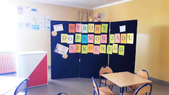 Sala lekcyjna. W&nbsp;centralnym punkcie ciemna ściana z&nbsp;kolorowym napisem "Wybory do samorządu szkolnego". Po lewej urna wyborcza.