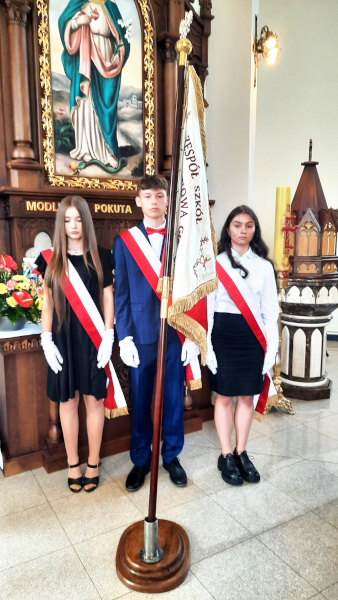 Troje uczniów wchodzących w&nbsp;skład pocztu sztandarowego stoi przy sztandarze szkoły wewnątrz kościoła, przed uroczystą mszą świętą na zakończenie roku szkolnego.