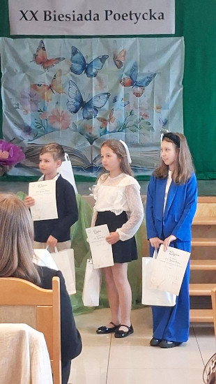 Trójka dzieci stoi na scenie podczas podsumowania XX Biesiady Poetyckiej, każda z nich trzyma dyplom i torbę upominkową.