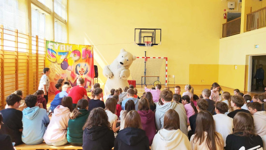 Dzieci siedzą w&nbsp;sali gimnastycznej, oglądając występ osoby w&nbsp;stroju niedźwiedzia polarnego, na tle kolorowego baneru „Cyrk Szok”.