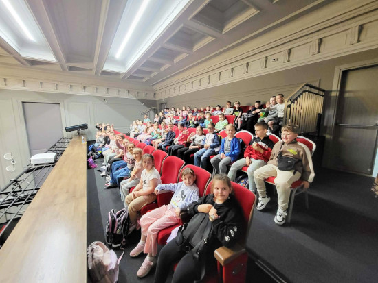 Grupa dzieci siedzi na widowni sali kinowej lub teatralnej, przygotowując się do oglądania spektaklu „Smocze Opowieści” w GCK.