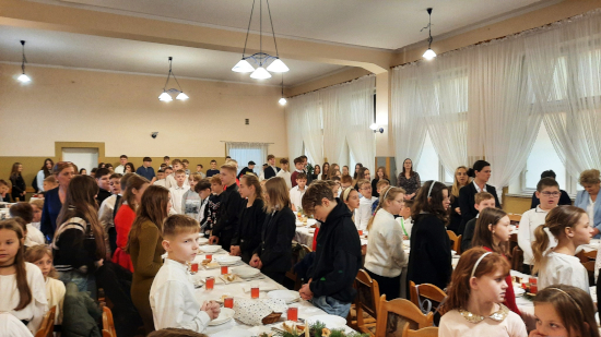 Uczniowie zgromadzeni w&nbsp;sali podczas szkolnej wigilii, stoją przy stołach nakrytych białymi obrusami i&nbsp;świątecznymi dekoracjami.