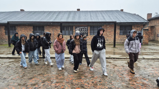 Grupa uczniów w&nbsp;ciepłych ubraniach idzie po mokrej, ceglanej nawierzchni na terenie dawnego obozu Auschwitz-Birkenau.