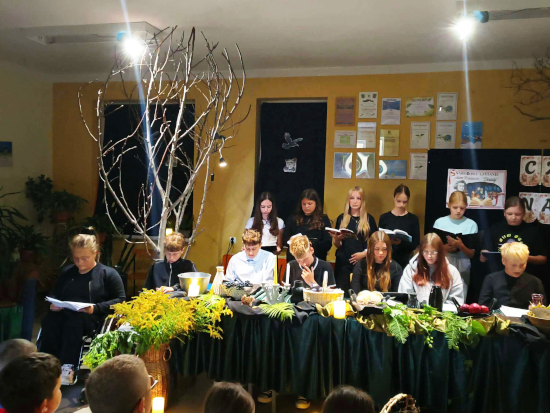 Grupa młodych osób czyta fragmenty dramatu „Dziady” przy udekorowanym stole z roślinami i lampionami, w sali oświetlonej reflektorami.